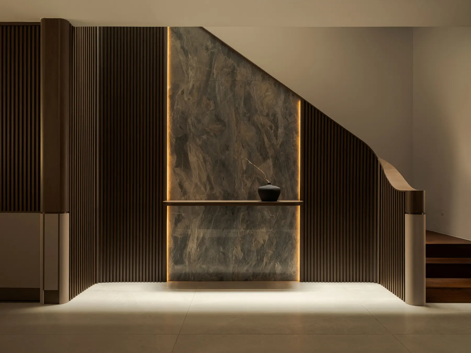 Staircase foyer — lit stone feature wall, vertical timber slats, ledge shelf with ceramic vessel