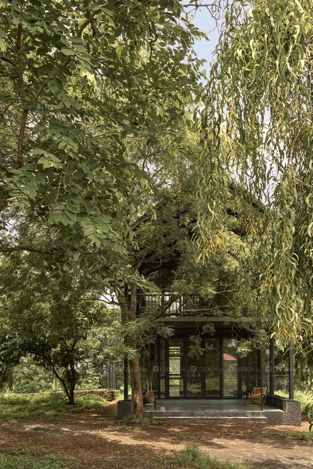 A glazed volume opens toward the established tree canopy, with a wooden chair on the verandah visible within